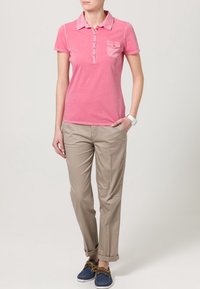 Polo shirt rosa a maniche corte con tasca e bottoni, abbinato a pantaloni beige chiaro; indossato con scarpe blu con lacci.