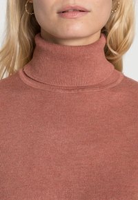Maglione a collo alto in tonalità ruggine con texture a costine. Il colletto alto e il design aderente aumentano calore e comfort. Nessun motivo o accento visibile.