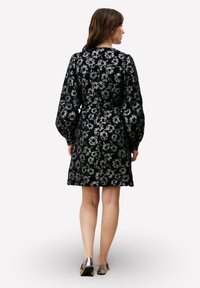 Zwarte bloemenjurk met een getextureerd ontwerp, voorzien van lange mouwen en een strakke taille. De stof heeft een subtiele glans en een ingewikkeld bloemmotief.