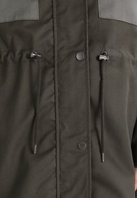 Mörkgrön jacka med slät tyg, knappstängningar, dragsko i midjan och en kontrasterande grå yoke-sektion.