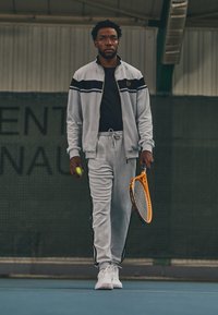 Grå treningsdrakt med svart striper, tennisball i den ene hånden, oransje tenniskølle i den andre, og hvite sneakers. Innendørs setting.