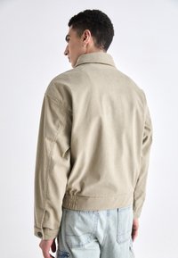 Beige kordfløyelsjakke med bred krage, senkede skuldre og elastisk nederkant; har knappede mansjetter og ribbet tekstur.
