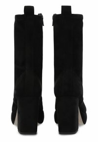 Un par de botas de tacón alto de ante negro hasta el tobillo con cremallera trasera y tiradores, vistas desde atrás sobre un fondo blanco.