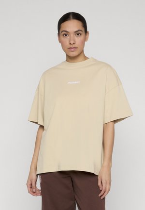 Young woman with dark hair tied back wears a beige oversized T-shirt with white text and brown pants, standing against a plain background.