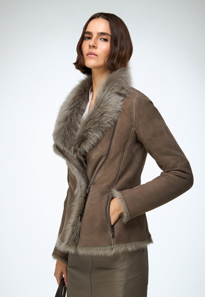 Veste en daim marron avec col et poignets en fourrure, dotée d'une fermeture éclair à l'avant. Le design comprend des poches latérales et une silhouette ajustée.