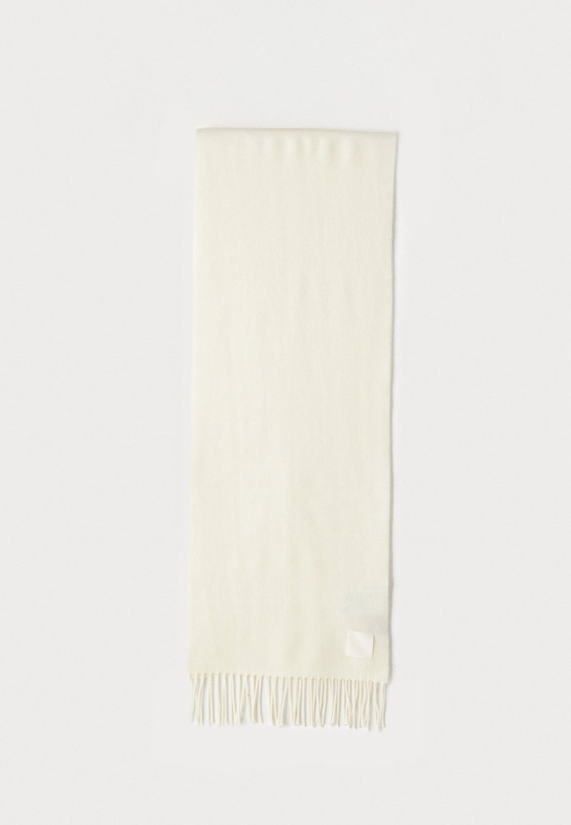 Écharpe en cream clair avec une texture douce, des franges aux extrémités et une petite étiquette en tissu dans le coin inférieur.