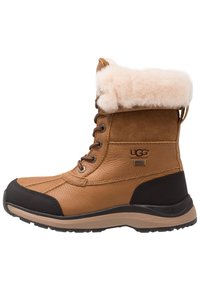 Bottes imperméables marron avec tige en daim et cuir, col crème duveteux, design à lacets, semelle extérieure en caoutchouc noir et logo de marque bien visible.