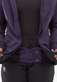 Veste imperméable violette avec fermeture éclair à l'avant et boutons-pression, associée à un pantalon noir. Tissu lisse avec des détails de coutures visibles.