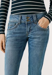 Jeans en denim bleu avec une coupe ajustée, comportant cinq poches, une fermeture à double bouton et des détails de surpiqûre contrastés. Lavage léger avec une légère texture.