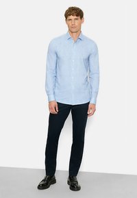 Hellblaues Hemd mit subtiler Textur, kombiniert mit dunkelblauen, schmal geschnittenen Hosen und schwarzen Schuhen. Einfaches Design, klassischer Schnitt.