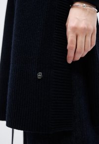 Μπλε ναυτικό πλεκτό πουλόβερ με ασημένες μεταλλικές λεπτομέρειες, με υφή υφάσματος και εφαρμοστή σχεδίαση. Χέρι ακουμπάει στο ένδυμα.