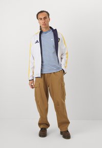 Chaqueta blanca con acentos en marine y franjas amarillas, superpuesta sobre una camisa de rayas azules, combinada con pantalones cargo beige y botas marrones.