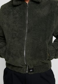 Groene fleecejas met een ritssluiting aan de voorkant, een hoge kraag en zakken in de zijnaden. Geweven stof met een zachte, knusse uitstraling.