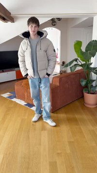 Beige puffer jacket with a hood, gray sweater, light blue jeans, and white sneakers. Interior features a brown ribbed couch and a plant.
