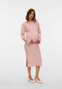 Jasnoróżowy, prążkowany zestaw swetra i spódnicy ciążowej z długimi rękawami, wysokim rozcięciem po boku spódnicy oraz białymi butami na słupku.