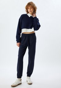 Navy cropped sweatshirt over een witte blouse, gecombineerd met navy sweatpants met elastische boorden en een trekkoord, afgewerkt met witte sneakers.