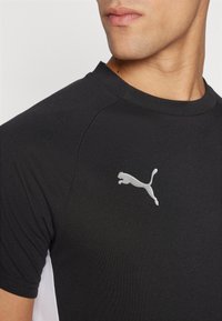 Tricou sportiv negru cu mâneci scurte, guler rotund și un logo Argintiu Puma în zona stânga a pieptului. Textură de țesătură netedă.