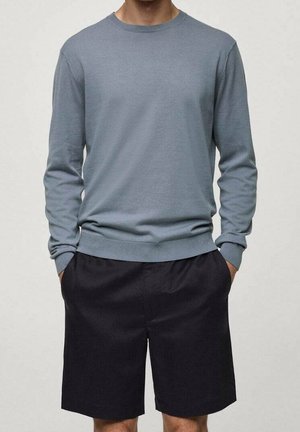 Person iført en grå langærmet sweater og sorte knælange shorts med hænderne i lommerne mod en ensfarvet lys baggrund.