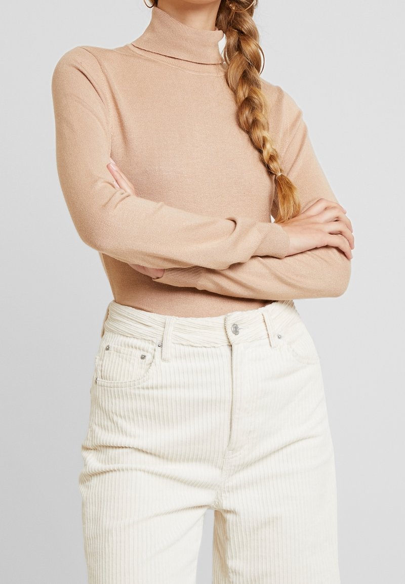 Femme aux cheveux tressés portant un pull à col roulé beige et un pantalon en velours côtelé blanc taille haute, bras croisés devant un fond uni.
