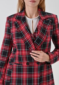 Karierte Blazer in Rot und Schwarz mit doppelter Knopfleiste, großen Revers, seitlichen Taschen und einer glatten Stofftextur. Darunter wird ein weißes Hemd getragen.
