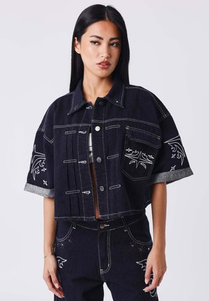 Mujer con chaqueta corta de denim oscuro y pantalones a juego con patrones bordados en blanco y mangas remangadas, mirando hacia un lado.