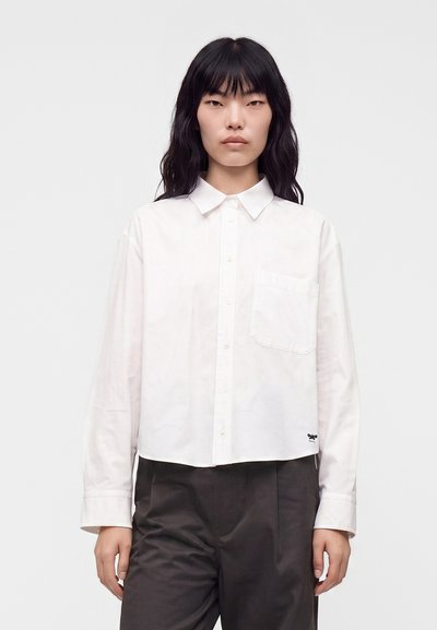 Femme aux longs cheveux noirs portant une chemise blanche boutonnée avec poche poitrine et un pantalon ample foncé, debout devant un fond uni.