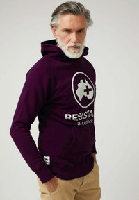 Sudadera con capucha morada con un bolsillo frontal. Presenta un gran logo blanco y el texto "RESISTENCIA" debajo. Fabricada con tela suave, con una apariencia texturizada.