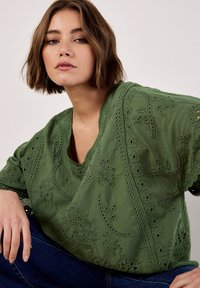 Grüne bestickte Bluse mit lockerer Passform, die mit Blumenmustern, Durchbrüchen und einem V-Ausschnitt versehen ist. Kombiniert mit dunkelblauen Jeans.