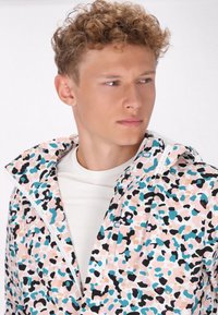 Farverig, letvægtsjakke med en pastelpink bund, sort og teal abstrakte pletter, lynlåsåbning og hætte.