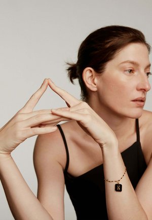 Mujer con el cabello oscuro recogido que lleva una blusa negra de tirantes y una pulsera con un charm negro marcado con "K", con los dedos tocándose para formar una forma de triángulo.