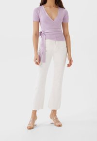 Kvinna som bär en lila omslagstopp med korta ärmar, vita cropped byxor och sandaler med genomskinlig klack på en vit bakgrund.