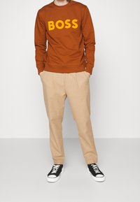 Sudadera marrón con "BOSS" destacado en amarillo. Combinada con pantalones plisados beige y zapatillas negras con suelas blancas.