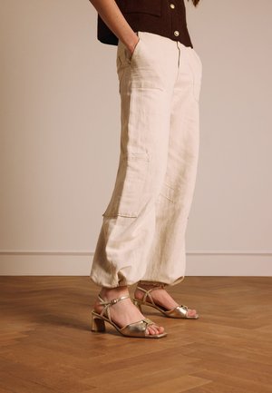 Persoon die beige losse cargobroeken en metallic gouden sandalen met lage hak draagt, staand op een houten parketvloer met handen in de zakken.