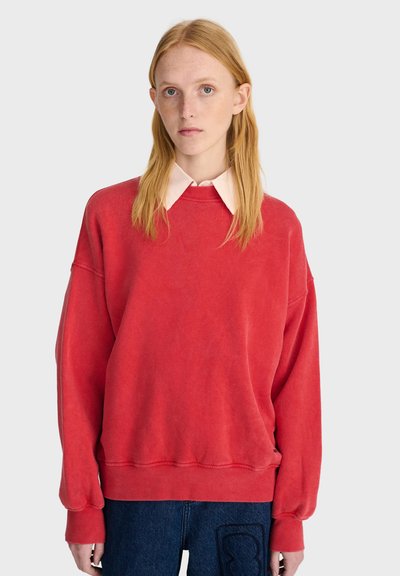 Jeune personne aux cheveux roux raides porte un sweat-shirt rouge oversize par-dessus une chemise blanche à col et un jean bleu sur un fond uni.