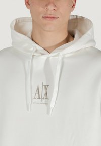 Balta kapučinė striukė su raišteliais, su išsiuvinėtu "AX" logotipu pilka spalva. Lygus paviršius, minimalistinio dizaino, pagaminta iš medvilnės.