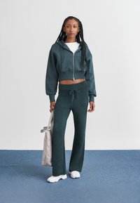 Kratek zip-up kapucar in udobne hlače temno zelene barve, z elastičnim pasom. Model drži svetlo sivo torbo in nosi bele čevlje.