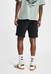 T-shirt surdimensionnée vert clair avec imprimé graphique, shorts noirs, chaussettes blanches avec chevrons noirs et baskets blanches avec accents noirs.