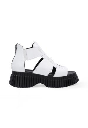 Hvide læder platform sandaler med en tyk sort ribbet sål, der har krydssnore-stropper og en lynlås lukning på siden.
