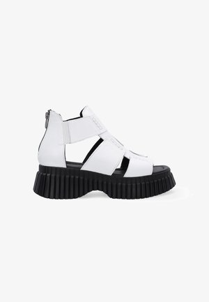 White leather platform sandals with a thick black ribbed sole, featuring crisscross straps and a side zipper closure.