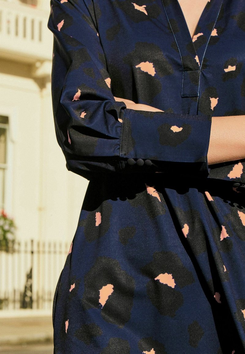 Personne portant une robe bleu foncé avec des taches abstraites noires et roses, bras croisés, debout à l'extérieur près d'un bâtiment clair.