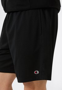 Shorts sportivi neri in tessuto rete, caratterizzati da una vestibilità ampia, tasche laterali e un piccolo logo rosso e bianco sul lato sinistro in basso.