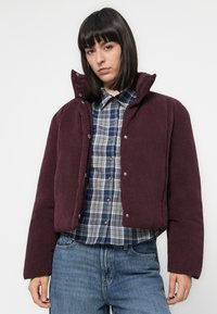 Jongere met rechtstreeks donker haar, gekleed in een paarse corduroy pufferjack over een marineblauw en wit geruit overhemd en blauwe spijkerbroek.