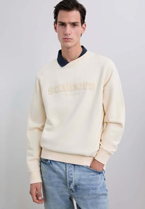 Joven vestido con un sudadera de escamas crema con el texto "scalpers" y vaqueros azul claro, de pie frente a un fondo gris claro.