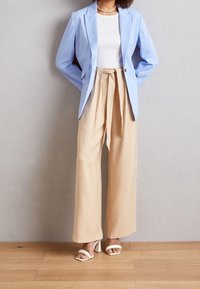 Lichtblauwe blazer, witte getailleerde top, beige hooggetailleerde broek met ceintuurlint en witte open teen hakken. Soepele stof, nauwsluitende pasvorm.