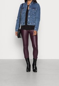 Woman standing against a neutral background, wearing a denim jacket, black top, burgundy leggings, and black heeled boots, holding a handbag.