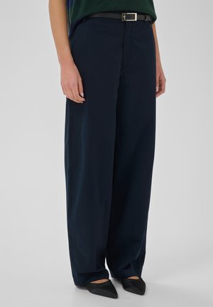 Persona che indossa pantaloni scuri a gamba larga, scarpe nere a punta e una cintura nera; la parte superiore del corpo è parzialmente visibile su uno sfondo semplice.