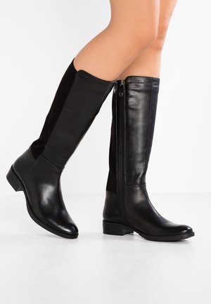 Beinlanger Blick auf eine Person, die kniehohe schwarze Lederstiefel mit seitlichen Reißverschlüssen trägt, stehend auf einer weißen Oberfläche vor weißem Hintergrund.