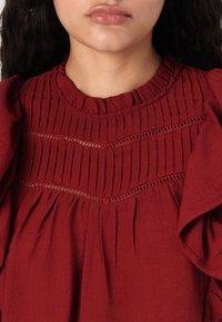 Blouse rouge avec un col texturé en volants et des manches bouffantes. Présente des plis verticaux et des découpes en dentelle sur la poitrine. Tissu doux.