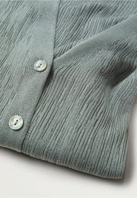 Tessuto di cardigan verde chiaro con texture e righe verticali, caratterizzato da due bottoni rotondi e lucidi di colore contrastante.