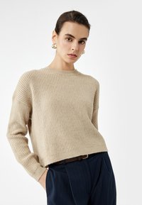 Beżowy sweter dzianinowy z okrągłym dekoltem, opadłymi ramionami i strukturalnym wzorem. Noszony na granatowe spodnie w paski.